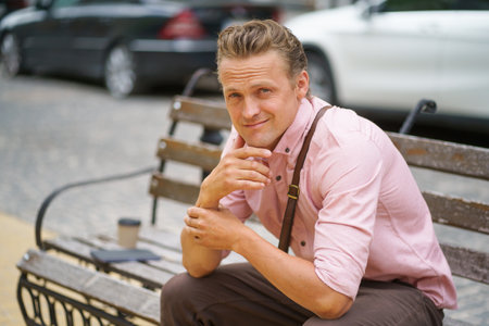 Handsome individual seated on a bench in the charming streets of an old town. With a warm and genuine smile, he confidently gazes at the camera, radiating charm and positive energy. . High quality photoの写真素材
