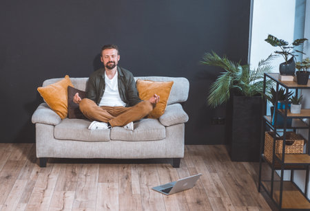Man sitting home on couch, adopting lotus asana pose, and embracing state of relaxation. With serene expression on face, he immerses himself in practice of meditation or mindfulness. High quality photoの写真素材