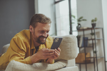 Happy and smiling man texts message on phone while lying on sofa at home. man enjoys convenience of modern technology in the cozy environment of his own home. . High quality photoの写真素材