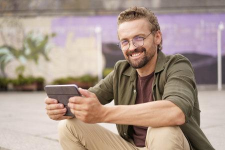 Mid-aged, handsome man sitting on the stairs of an embankment, engrossed in reading good news on his tablet PC. A genuine smile lights up his face as he discovers positive and uplifting information. . High quality photoの写真素材