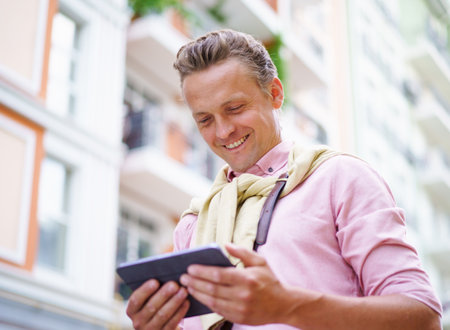 Happy man reading latest news articles from tablet PC while being outdoors on city street. Tablet PC in his hands allows him to access the latest information and stay up-to-date with current events. . High quality photoの写真素材