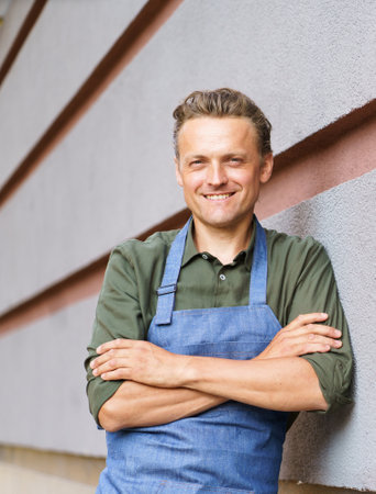 Skilled and dedicated waiter or service man in restaurant setting. Dressed in neat apron and uniform, he stands by the wall with crossed hands, radiating confidence and professionalism. . High quality photoの写真素材