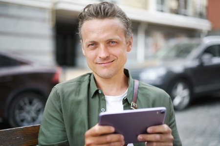 Texting message concept as man smiles while holding tablet PC on bench outdoors. With joyful expression on face, he engages in digital communication and stays connected in urban environment. High quality photoの写真素材