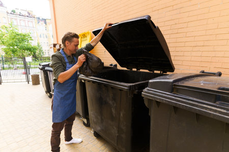 Waiter or kitchen worker responsibly disposing of garbage in pocket and placing it in city trash bin located in back yard. Focus on maintaining cleanliness and proper waste management in urban environment. . High quality photoの写真素材