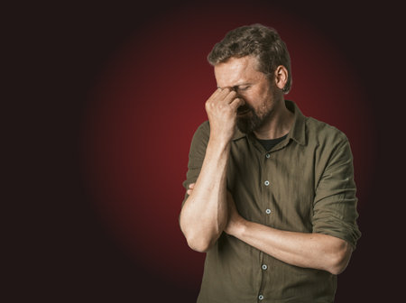 Man facing personal problems and challenges. Mid-aged man with closed eyes, conveying sense of sadness and inner turmoil. Isolated on dark red background, feeling of despair and emotional struggle. . High quality photoの写真素材