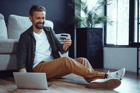 Happy customer is depicted sitting on floor at home with laptop, engaged in online payment process. Holding credit card, customer enters necessary data to complete online purchase. . High quality photoの写真素材