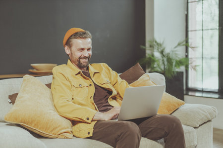Smiling young man enjoys comfort of home while working on laptop. Laptop on knees, exemplifies modern lifestyle of remote work and telecommuting, showcasing perfect balance between work and relaxationの写真素材
