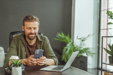 Moment in the office where a man is taking a break from working on the computer. He is seen texting a message on his phone, showcasing the multitasking nature of modern work environments. High quality photoの写真素材