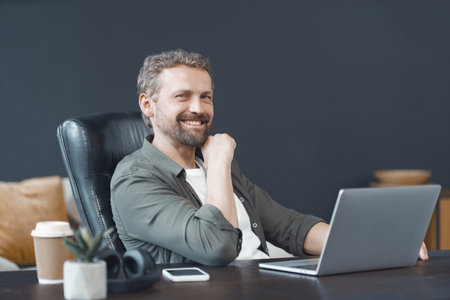 Smiling man finds joy in work as he diligently operates laptop on well-organized desk within bustling office environment. Corporate world, where he excels in his career with sense of fulfillment. High quality photoの写真素材