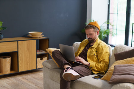 Tired man seeks solace on sofa home, battling exhaustion as engages with laptop, either in search of information or exploring websites on web. Modern digital age. High quality photoの写真素材