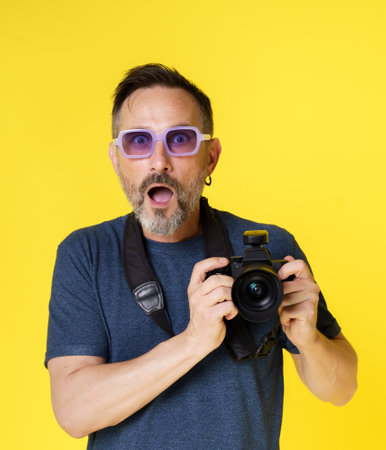 Passionate old photographer beams with excitement, holding mirrorless photo camera, in isolated studio shot on yellow background. Seasoned photographer continues pursue photography passion with zeal. High quality photoの写真素材