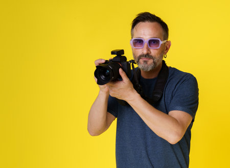 Stylish and creative mid-aged photographer with mirrorless camera against yellow background. Purple glasses adding touch of flair, modern and artistic photographer who involved both style and creativity. High quality photoの写真素材