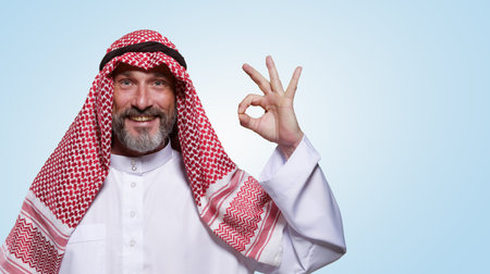Cheerful and confident Arab man smiles warmly and shows OK sign against light blue background, leaving space for text. Approval, and contentment, making versatile choice for various messaging needs. High quality photoの写真素材