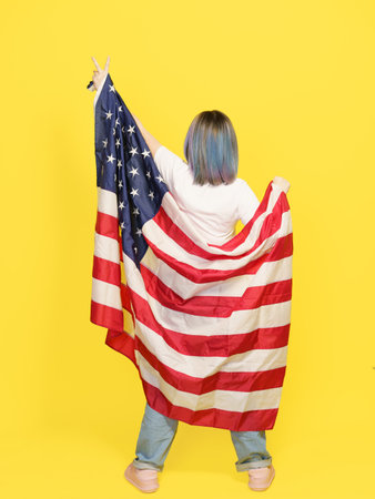 Victory Sign By Girl Covered With American Flag, Patriotic Pride, Cultural Diversity, And Freedom. Teenager Isolated On Yellow Backdrop With Copy Space For Your Text. Full-Height Backside View . High quality photoの写真素材