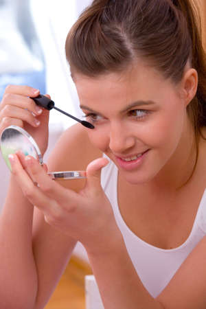 Young girl applying mascara on her eyelash and looking into a mirror