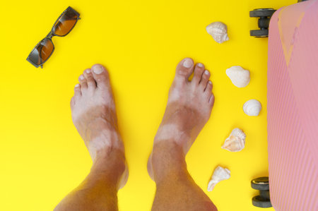 Tanned man's feet with seashells and suitcase on yellow background. Vacation cardの写真素材
