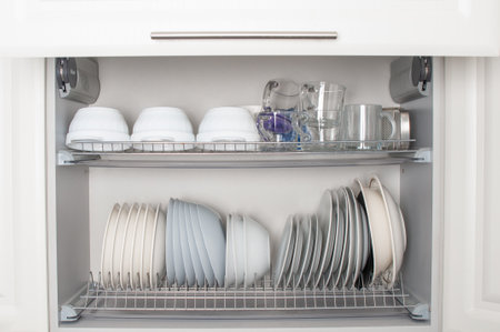 Clean dishes, mugs and different glasses in opened white kitchen cupboard shelfの写真素材