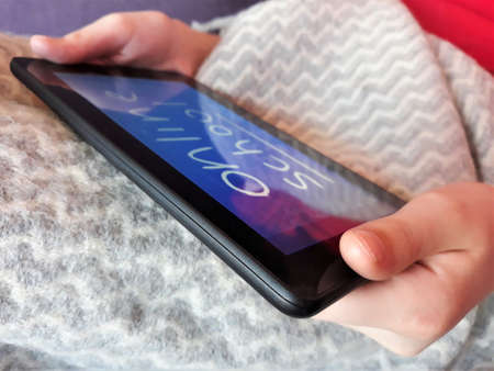 A tablet in the hands of a child with the text online school on the background of a gray plaid ckose-up. The concept of distance learning, online learning at home during the coronavirus epidemic, COVID-19.の写真素材