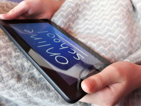 A tablet in the hands of a child with the text online school on the background of a gray plaid ckose-up. The concept of distance learning, online learning at home during the coronavirus epidemic, COVID-19.の写真素材