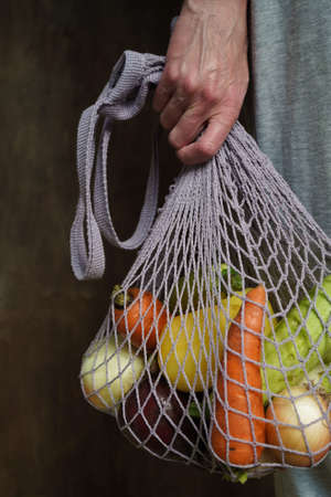 Woman holds her hand gray bag string bag with vegetables, zucchini, bell peppers, carrots, beets, onions brown wooden background.Harvest from garden fresh bright vegetables.Concept zero waste.の写真素材