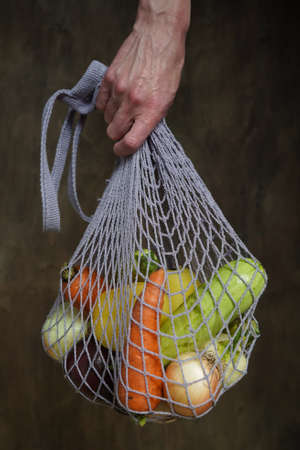 Woman holds her hand gray bag string bag with vegetables, zucchini, bell peppers, carrots, beets, onions brown wooden background.Harvest from garden fresh bright vegetables.Concept zero waste.の写真素材