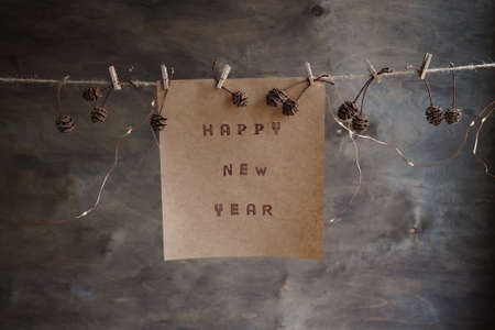 Happy new year text written on brown paper. New year poster hanging on a rope with clothespins and pine cones on a brown wooden background. New Year card conceptの写真素材