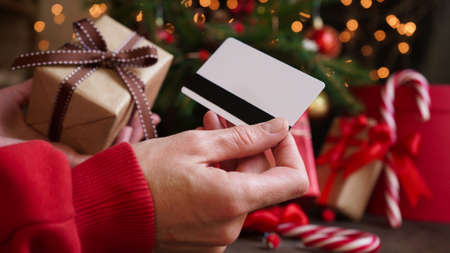 Young woman holding a credit card and a gift box against the background of Christmas decor and gifts, close-up. Christmas and New Year shopping on the Internet, payment by credit card.の写真素材
