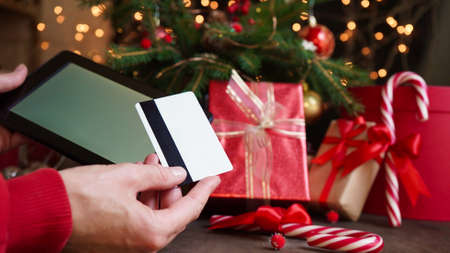 Young woman holding a credit card and a tablet the background of Christmas decor and gifts, close-up. Christmas and New Year shopping on the Internet, payment by credit card.の写真素材