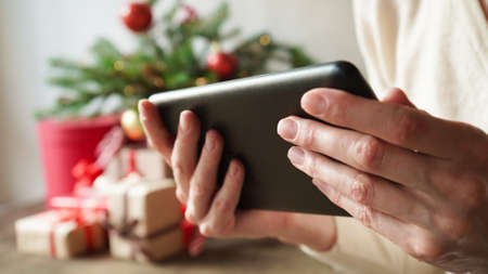 Young woman holding in hands a tablet the background of Christmas decor and gifts, close-up....