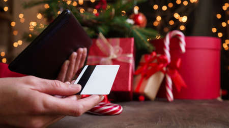 Young woman holding a credit card and a tablet the background of Christmas decor and gifts, close-up. Christmas and New Year shopping on the Internet, payment by credit card.の写真素材
