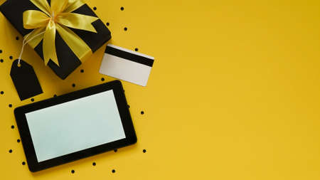 Online shopping for Black Friday and Christmas sales, pay by credit card. Black gift box with yellow bow, tablet, black tags, credit card, confetti on yellow background, top view, flat lay, copy space, close-up.の写真素材