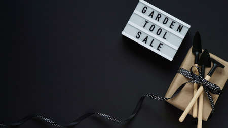 Banner concept for Black Friday celebration, lettering.White board with text GARDEN TOOL SALE, gift box with black pea bow and garden tools on black background, top view, flat lay, copy spaceの写真素材