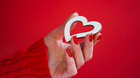 Close-up of female hands with red manicure holding white wooden heart on red background, copy space. Valentine's day concept. beautiful holiday card. love. Winter. Romanceの写真素材