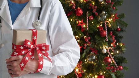 Concept for christmas and new year medical banner.Female doctor in white coat,stethoscope holds gift box with red bow in her hands against background beautifully decorated Christmas tree.Copy space.の写真素材
