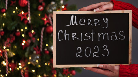 Marry christmas banner concept. Female hands in red sweater hold chalk board with text Merry Christmas 2023 on background of beautiful festive Christmas tree decorated with red toys and garlands.の写真素材