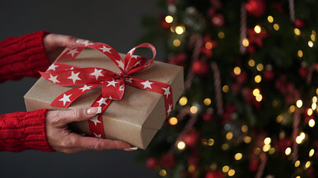 Female hands in red sweater hold Christmas New Year gift box on background of Christmas tree.Giving gifts for holidays.Christmas holidays celebration concept,gifts for family,friends.Winter banner.の写真素材