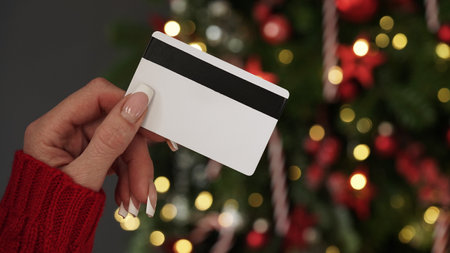 Hands of a woman in a red sweater with a credit card on the background of a Christmas tree close-up. Christmas and New Year shopping online, credit card payment. Buying gifts onlineの写真素材