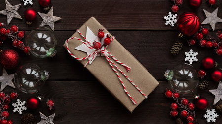 Gift wrapped in craft brown paper lies among holiday decor, red balls, stars, snowflakes and berries on wooden background, copy space, flat lay, top view.Merry Christmas,Happy New Year concept bannerの写真素材