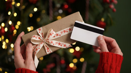 Hands of a woman in a red sweater holding a credit card and a gift box on the background of a Christmas tree close-up. Christmas and New Year shopping online, credit card payment. Buying gifts onlineの写真素材