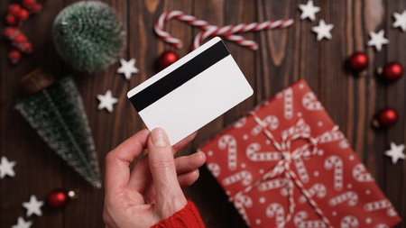 Hands of a woman in a red sweater with a credit card on a background of gift boxes, selective focus, close-up. Christmas and New Year shopping online, payment by credit card. Buying gifts onlineの写真素材
