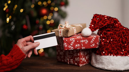 Hand of woman in red sweater holding credit card next to lot of gift boxes and santa claus hat against background of christmas tree.Christmas and New Year online shopping,payment by credit cardの写真素材