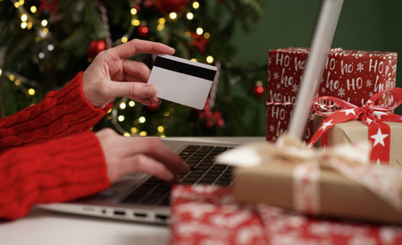 Young woman holding a credit card and makes purchases on a laptop against the background of Christmas decor and gifts, close-up. Christmas and New Years online shopping, credit card payments.の写真素材