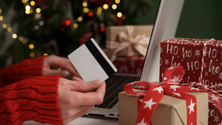 Young woman holding a credit card and makes purchases on a laptop against the background of Christmas decor and gifts, close-up. Christmas and New Years online shopping, credit card payments.の写真素材