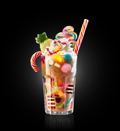 Monster shake, freak caramel shake isolated. Colourful, festive milk shake cocktail with sweets, jelly. Colored caramel milkshake array of different childs sweets and treats in glass on black background. Sweet candy milkshake. Crazy freakshake food trend.の写真素材