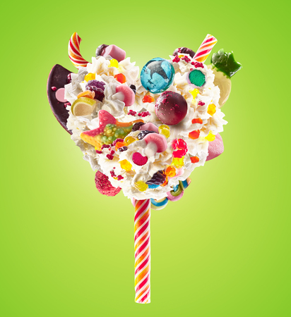 Sweet Lolipop in Heart form of whipped cream with sweets, jellies, heart front view. Crazy freakshake food trend. Front view of whipped heart of cream lolly, full of berry and jelly sweets, chocolate candy. Colored whipped heart form cream concept.の写真素材