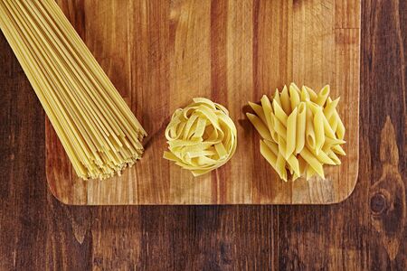 Various types of pasta on wooden table with cutting board, top view. Pappardelle, spaghetti, Penne pasta on dark wood table.の写真素材