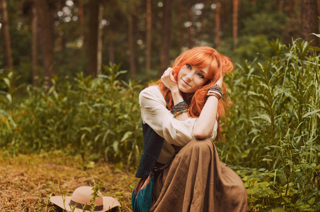 Redhead hippie walks in summer forest. Young woman with long hair in a long skirt and blouse in Boho style. Photo of romantic girl in national dress.の写真素材