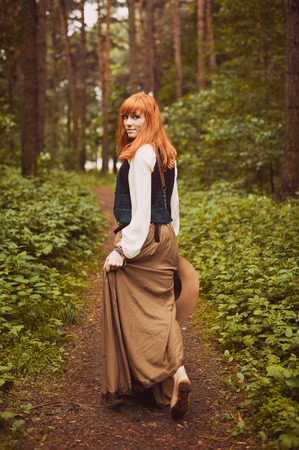 Redhead hippie walks in summer forest. Young woman with long hair in a long skirt and blouse in Boho style. Photo of romantic girl in national dress.の写真素材