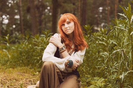 Redhead hippie walks in summer forest. Young woman with long hair in a long skirt and blouse in Boho style. Photo of romantic girl in national dress.の写真素材