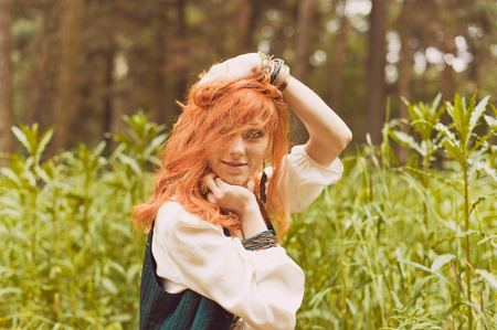 Redhead hippie walks in summer forest. Young woman with long hair in a long skirt and blouse in Boho style. Photo of romantic girl in national dress.の写真素材